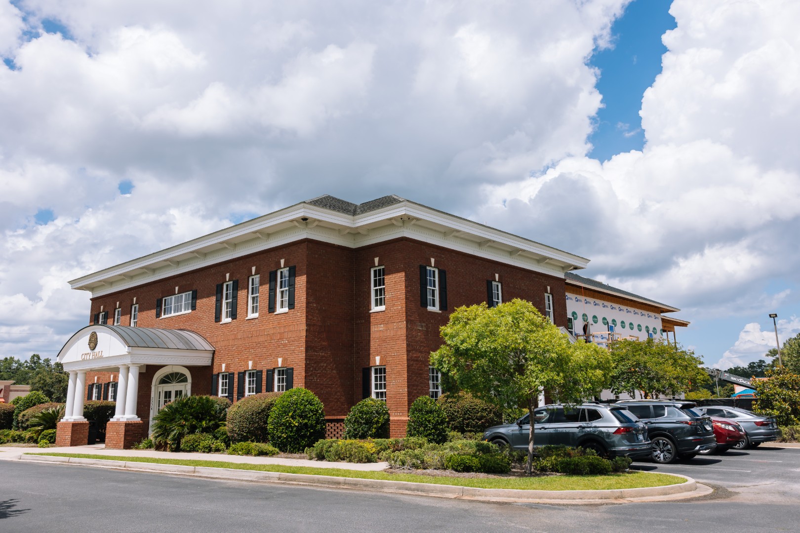 A two-story red brick building with white trim, surrounded by green bushes and trees, stands next to a parking lot filled with cars under a partly cloudy sky.