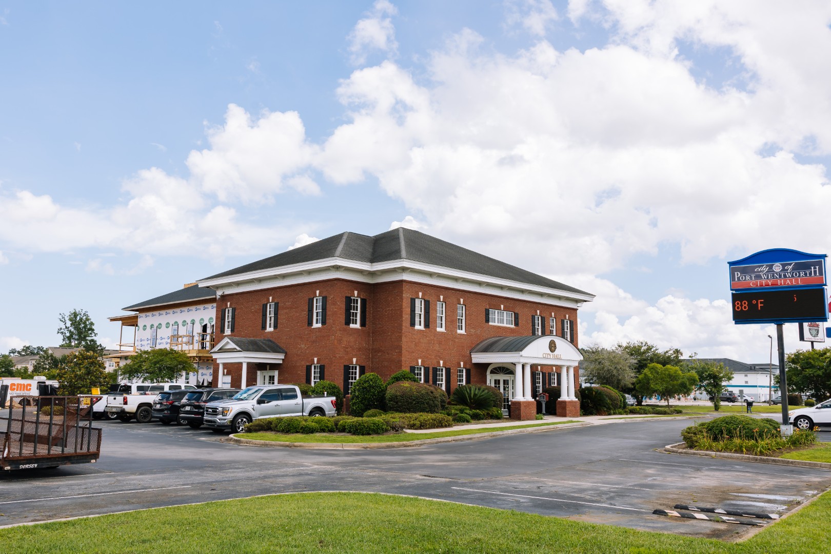 A two-story brick office building with white columns and arched windows, surrounded by parked cars and greenery under a partly cloudy sky.