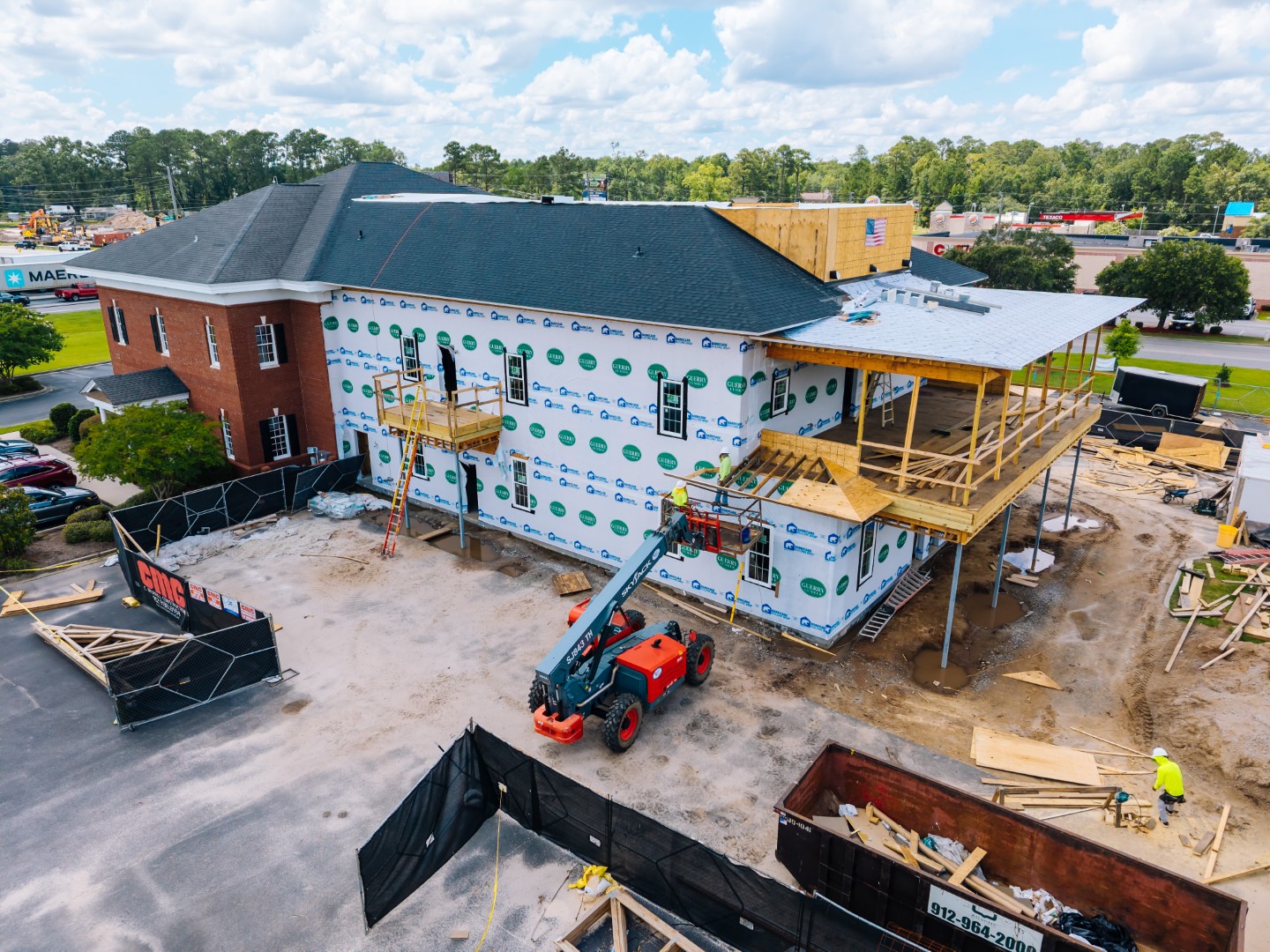 Aerial view of a building under construction, with workers, scaffolding, a forklift, and building materials around the site. The buildings exterior is partially covered in housewrap and the roof is mostly finished.