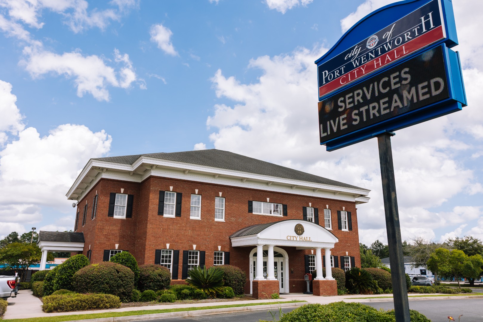 A red brick building with columns labeled City Hall is shown under a blue sky. In front, a digital sign reads, City of Port Wentworth City Hall.