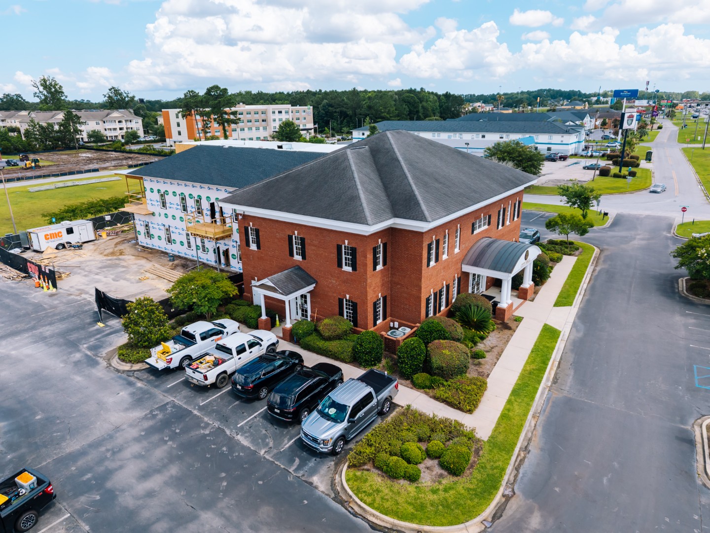 Aerial view of a two-story red brick office building with black roof, surrounded by neatly trimmed bushes and parked vehicles. Nearby, a building under construction is visible, along with a wide road and other structures in the background.