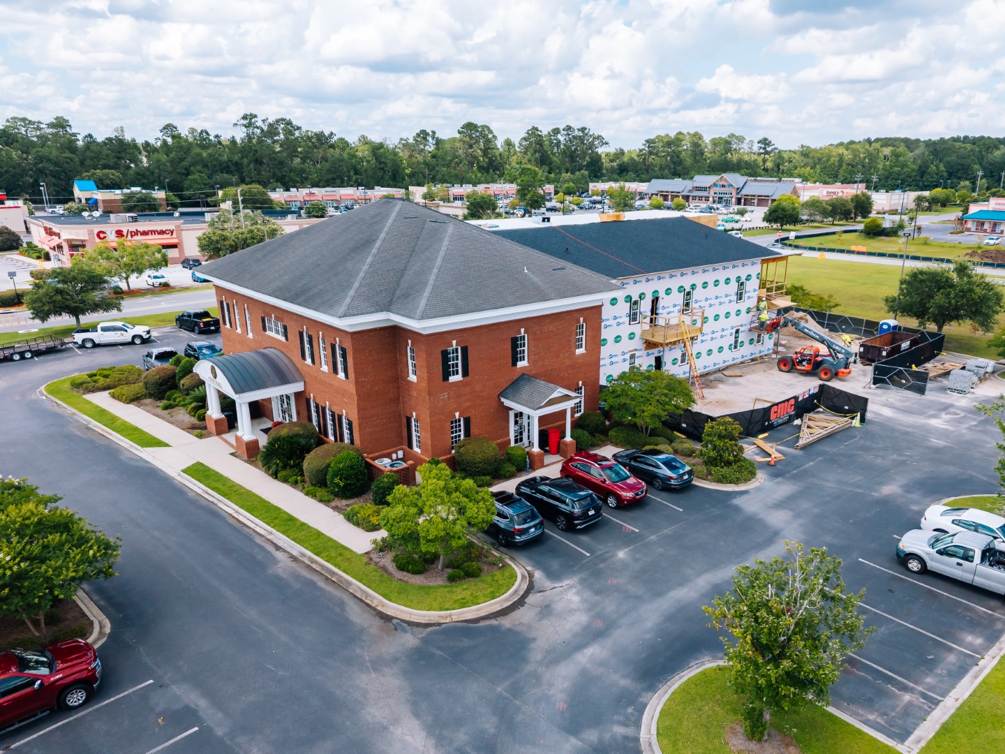 Aerial view of a brick office building with a parking lot and several cars; adjacent to it, a new structure is under construction with insulation visible. Surrounding area includes trees and nearby businesses.