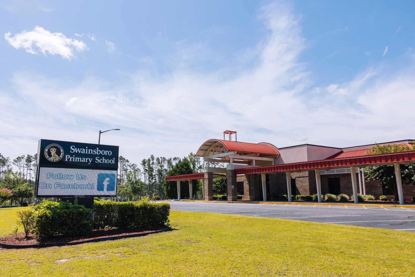 A building with a red roof and arched entrance, labeled “Swainsboro Primary School.” In the foreground, a sign reads “Follow Us On Facebook!” Grass and trees surround the school under a partly cloudy sky.