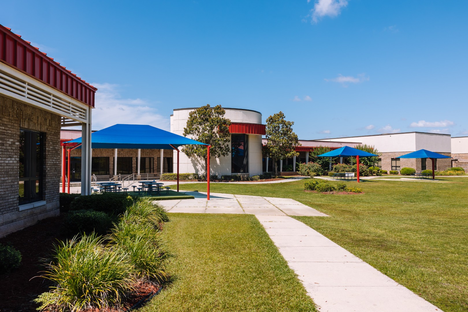 A school courtyard with green grass, trees, and blue canopy-covered picnic tables. The buildings have a mix of brick and white walls with red trim, under a clear blue sky.