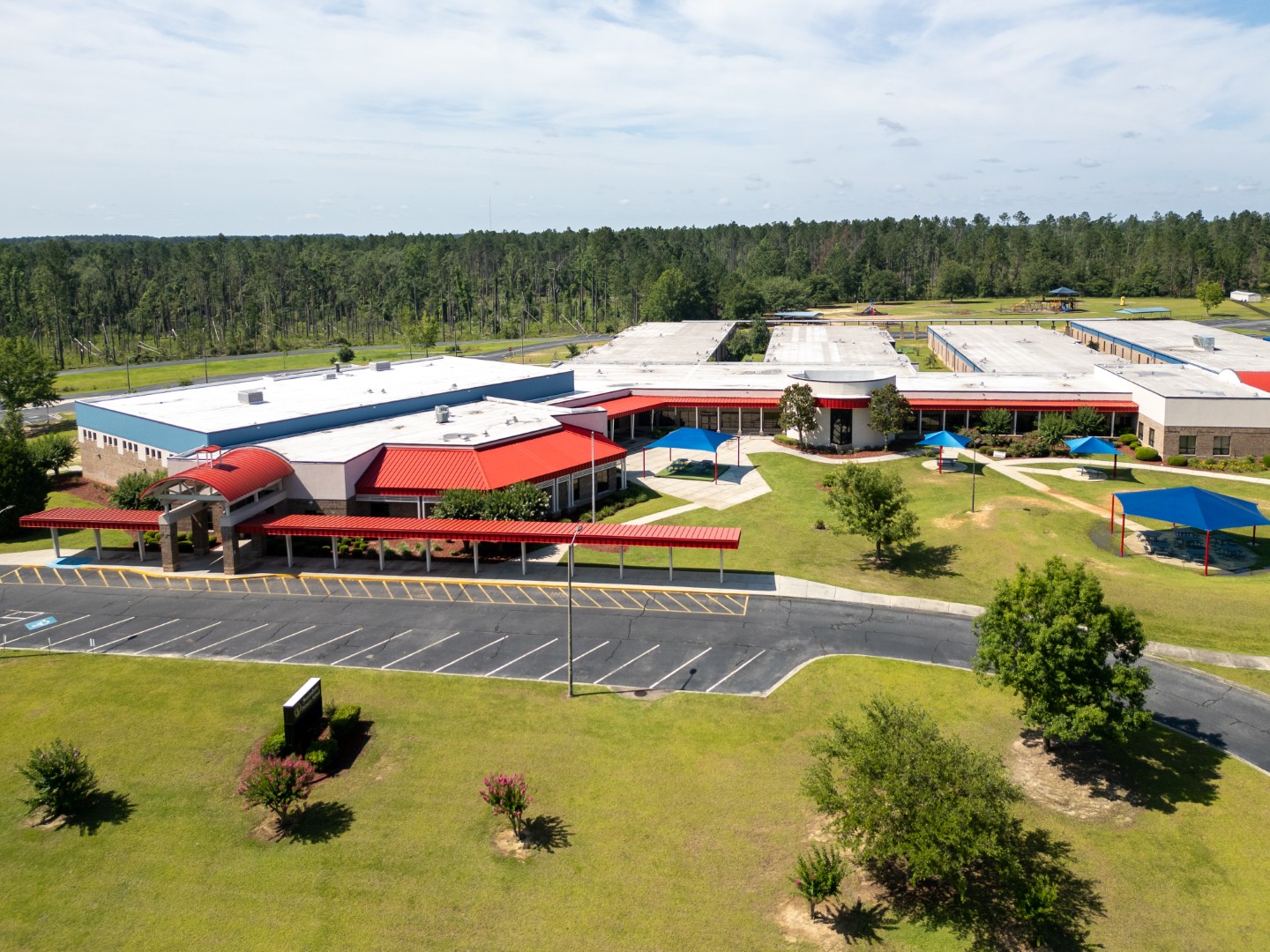 Aerial view of a large school campus with red and white roofs, surrounded by grassy fields, trees, and a few parking spaces. Covered walkways and outdoor seating areas are visible. Forested area lies beyond the campus.