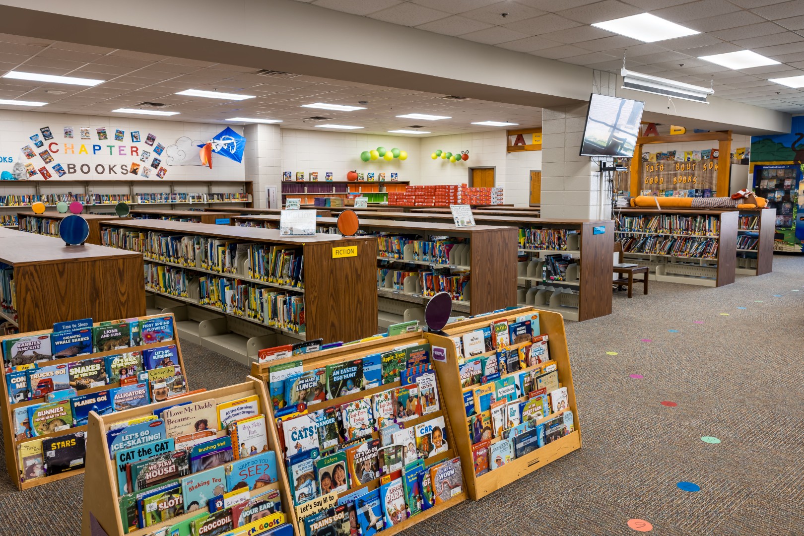 A spacious, well-lit library with bookshelves filled with children’s books, a section labeled “Chapter Books,” colorful signs, and a cozy reading area. The carpet has colorful dots, and wall decorations add a playful touch.