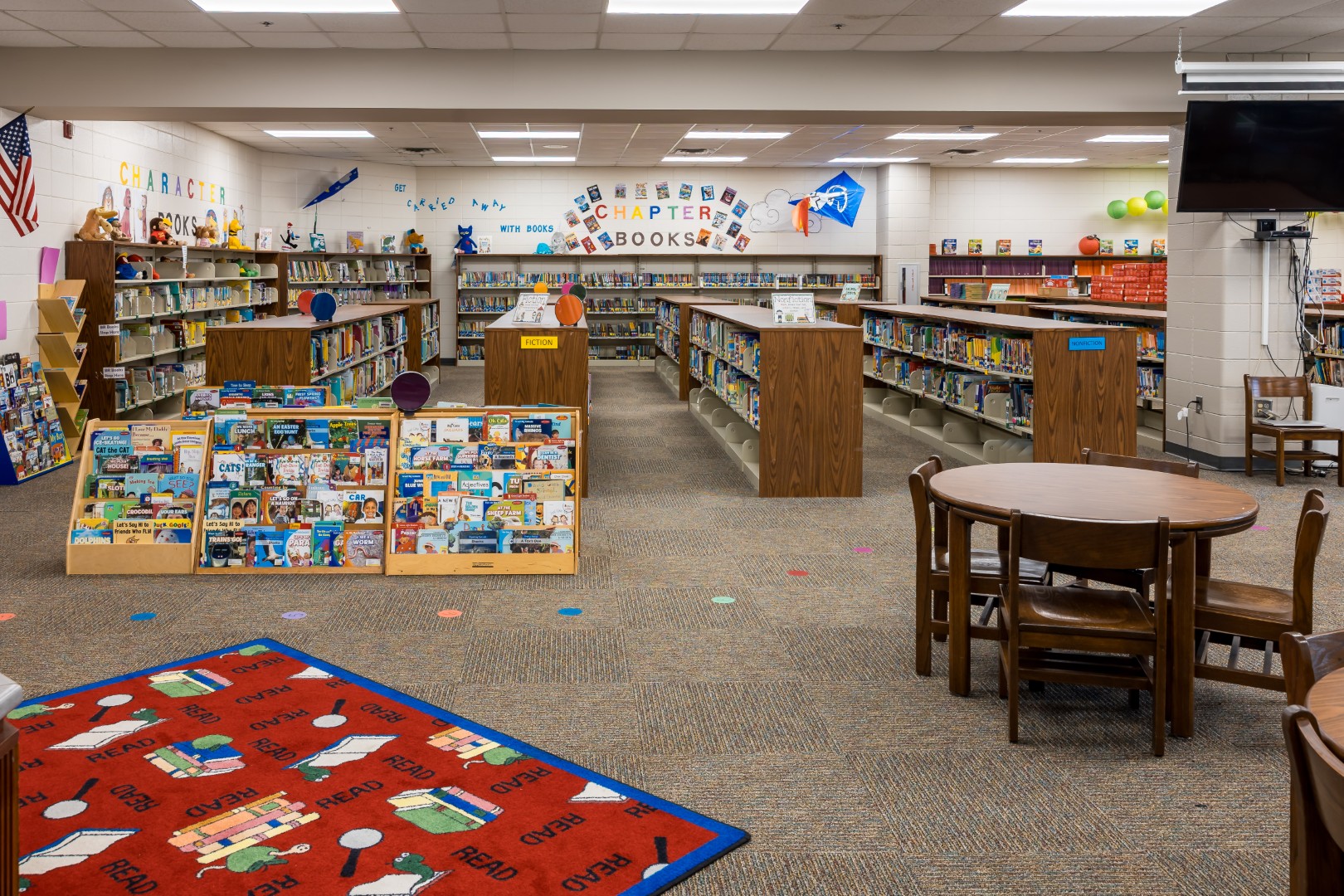 A brightly lit library with wooden bookshelves filled with books, a colorful rug that says READ, small round tables with chairs, and wall decorations including the words CHAPTER BOOKS.