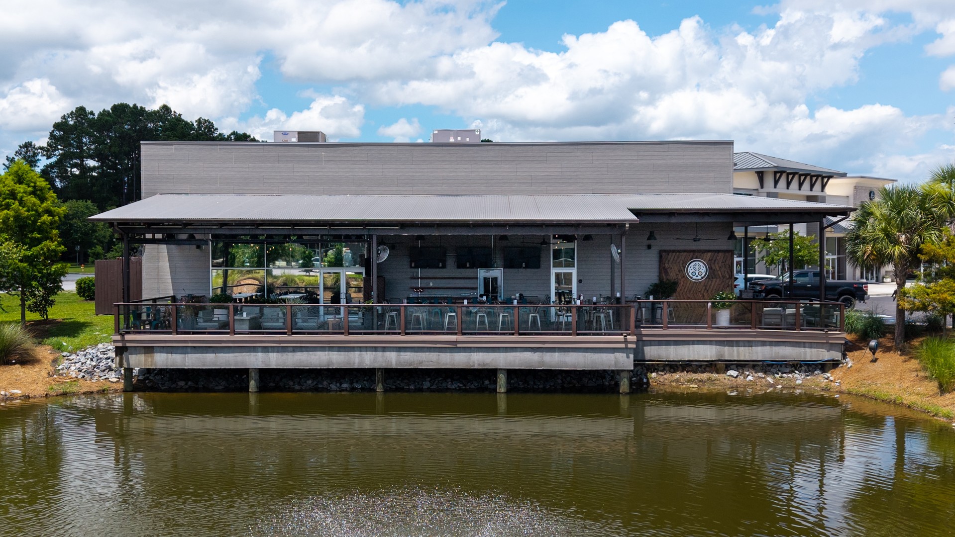 A gray lakeside restaurant with a covered outdoor seating area, large windows, and bar stools overlooks a pond. Trees and greenery surround the building under a partly cloudy sky.