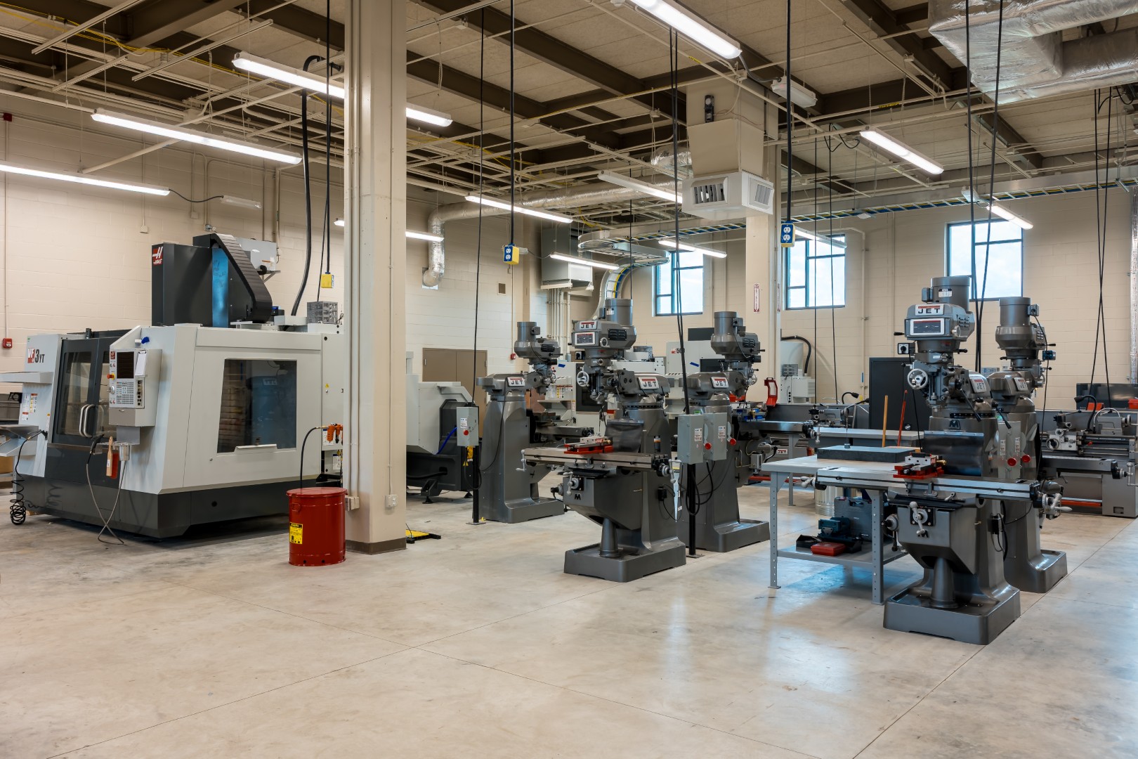 A spacious, well-lit machine shop with multiple milling machines, a large CNC machine, workbenches, and safety equipment arranged on a clean concrete floor. High ceilings and windows provide natural light.