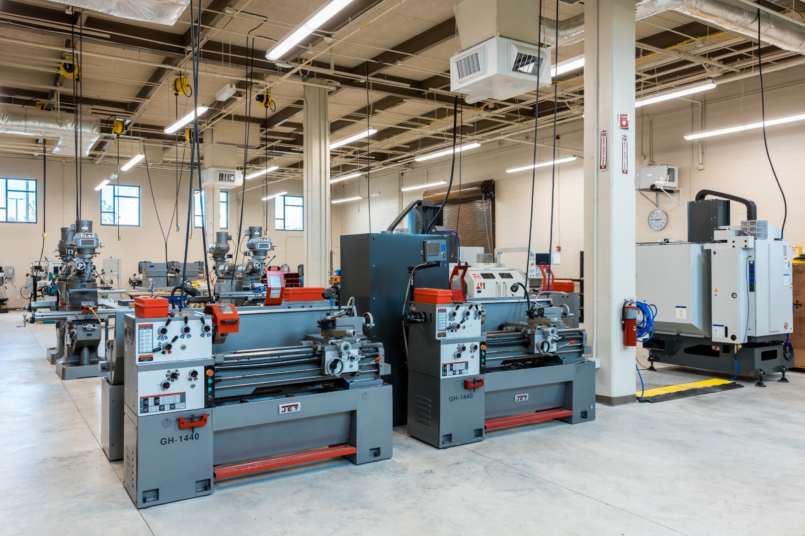 A spacious, well-lit workshop with multiple industrial lathes and milling machines arranged in rows on a polished concrete floor. The walls are light-colored, and large windows provide ample natural light.