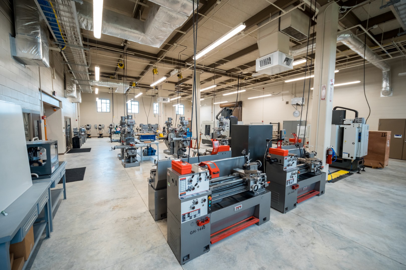 A spacious, well-lit industrial workshop with several metal lathes, milling machines, and other machining equipment arranged on a polished concrete floor. The room has high ceilings and exposed ductwork.