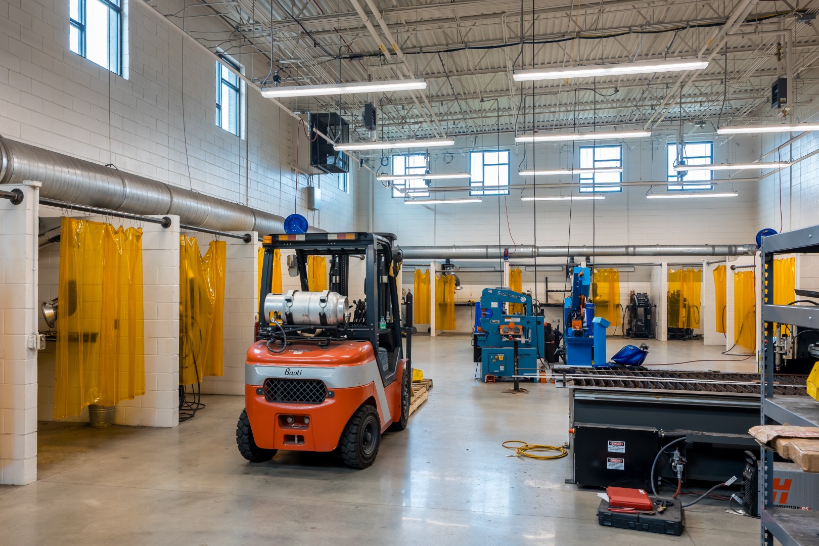A modern industrial workshop with a forklift in the center, yellow welding curtains, machinery, workstations, and large windows letting in natural light.