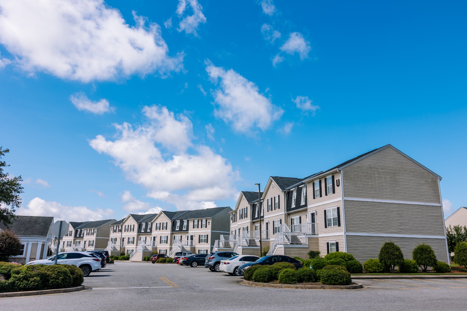A row of modern, light-colored apartment buildings with parked cars in front, set under a bright blue sky with scattered clouds.