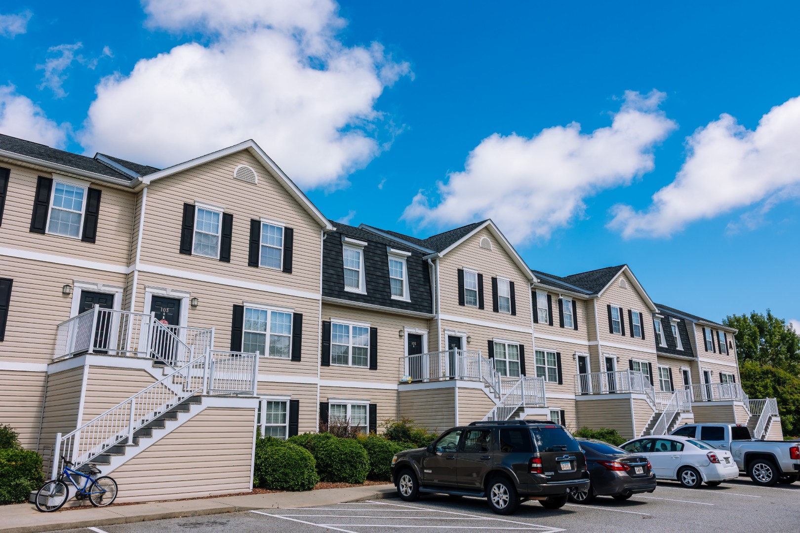 A row of beige, three-story apartment buildings with white railings and black shutters under a bright blue sky; several cars and a bicycle are parked in the lot in front.