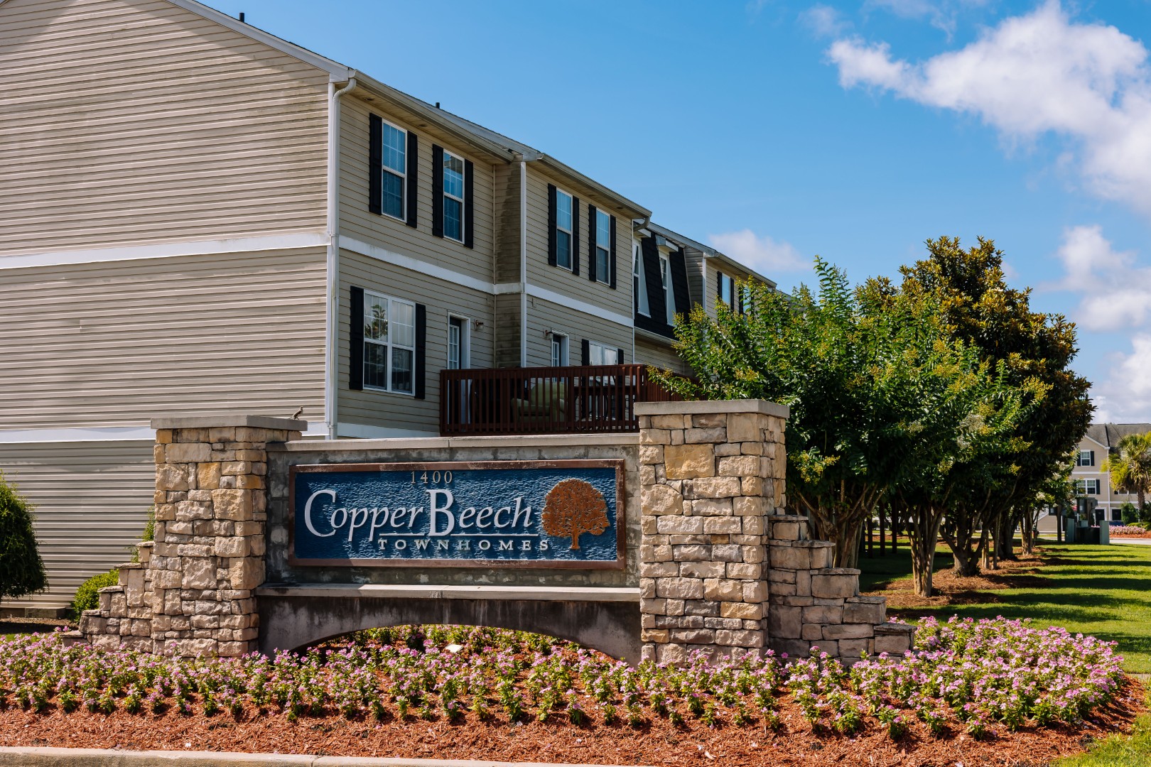 A sign reading Copper Beech Townhomes stands in front of tan townhouses with white trim, surrounded by landscaped flowers and greenery under a sunny blue sky.