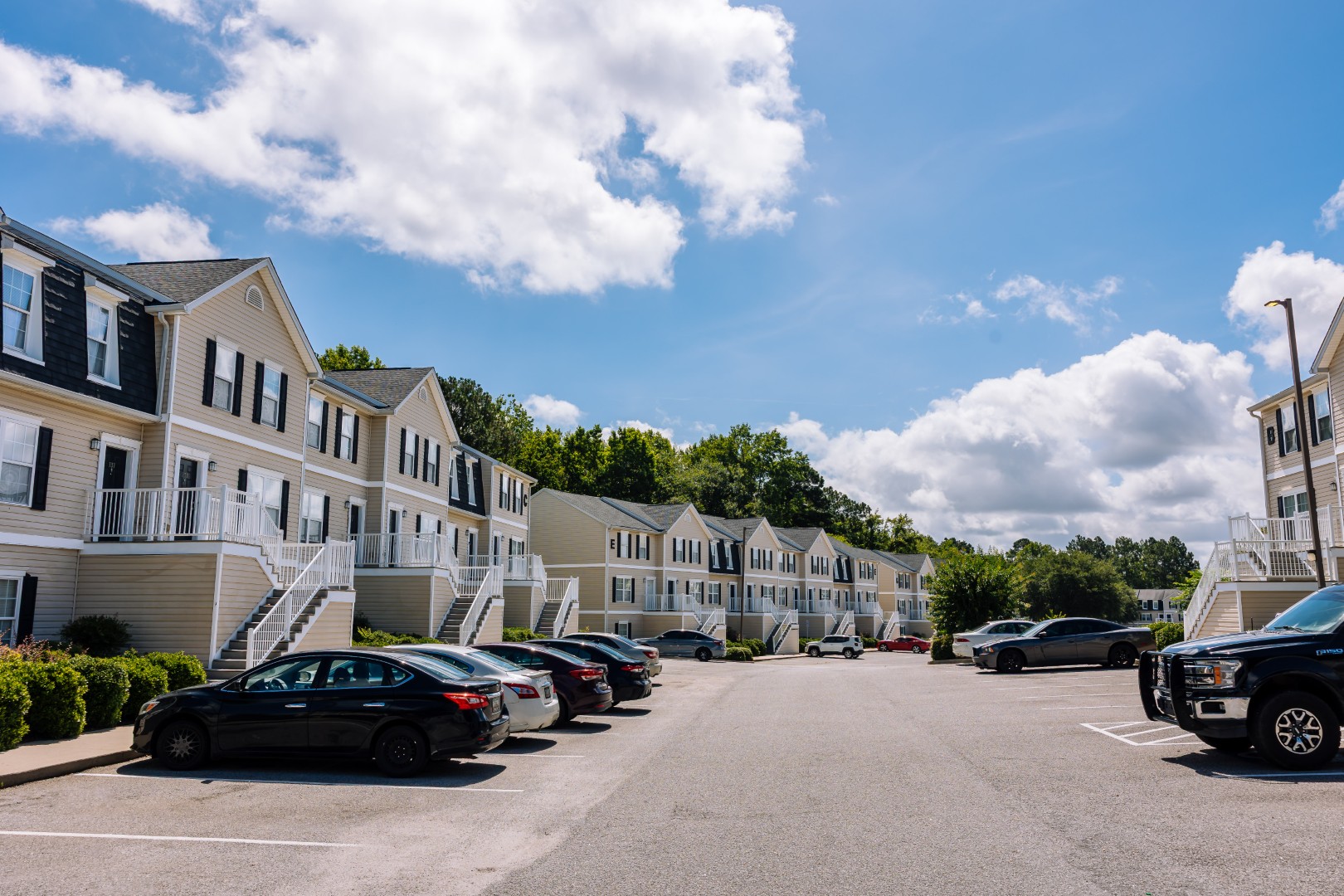 A wide view of a sunny apartment complex with multiple beige buildings, white trim, and parked cars lining the parking lot under a blue sky with scattered clouds.