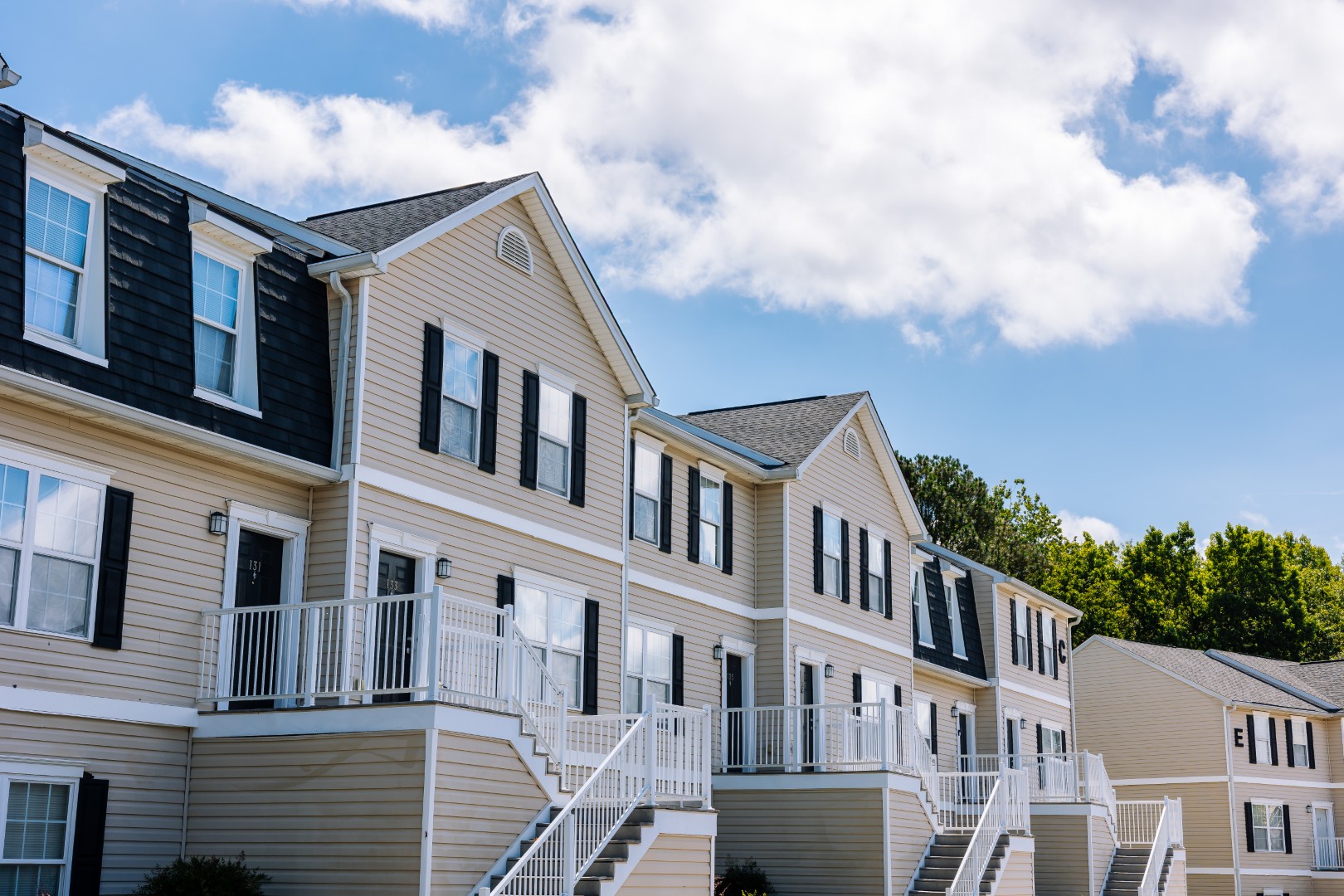 A row of modern beige townhouses with white staircases and railings, black shutters, and sloped roofs under a blue sky with scattered clouds and trees in the background.