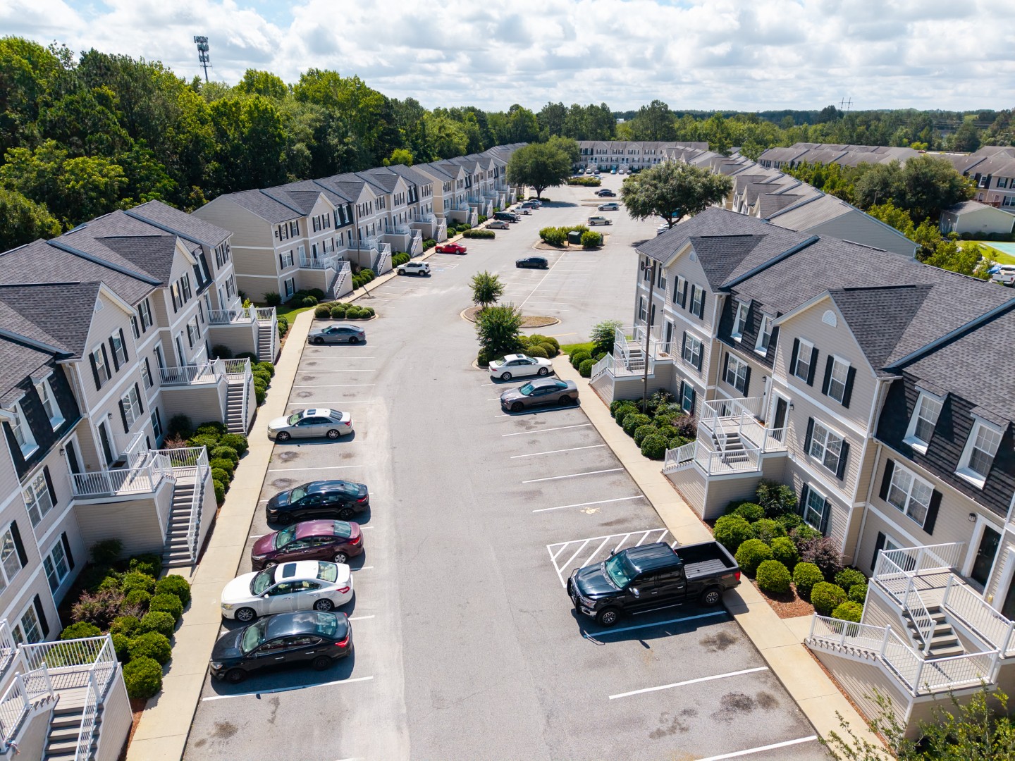 Aerial view of a residential apartment complex with rows of townhouses on both sides of a wide parking lot filled with parked cars, surrounded by greenery and trees under a partly cloudy sky.