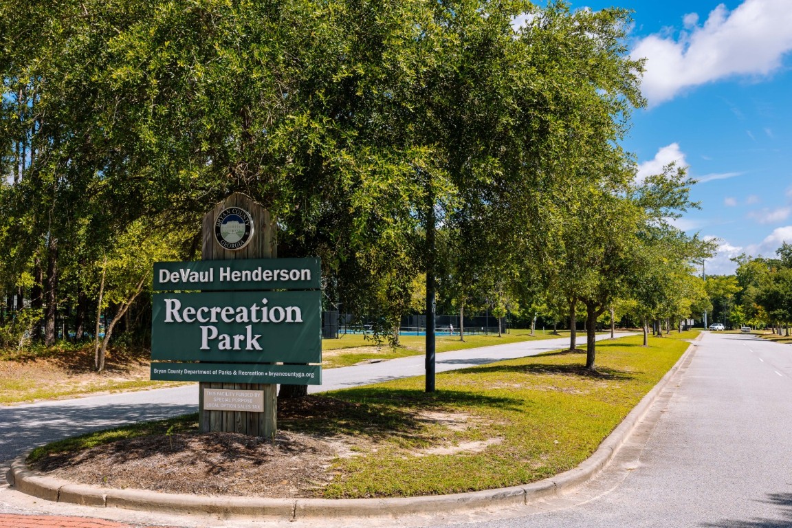 A large green sign reading DeVaul Henderson Recreation Park stands by a tree-lined road under a blue sky, with tennis courts and park facilities visible in the background.