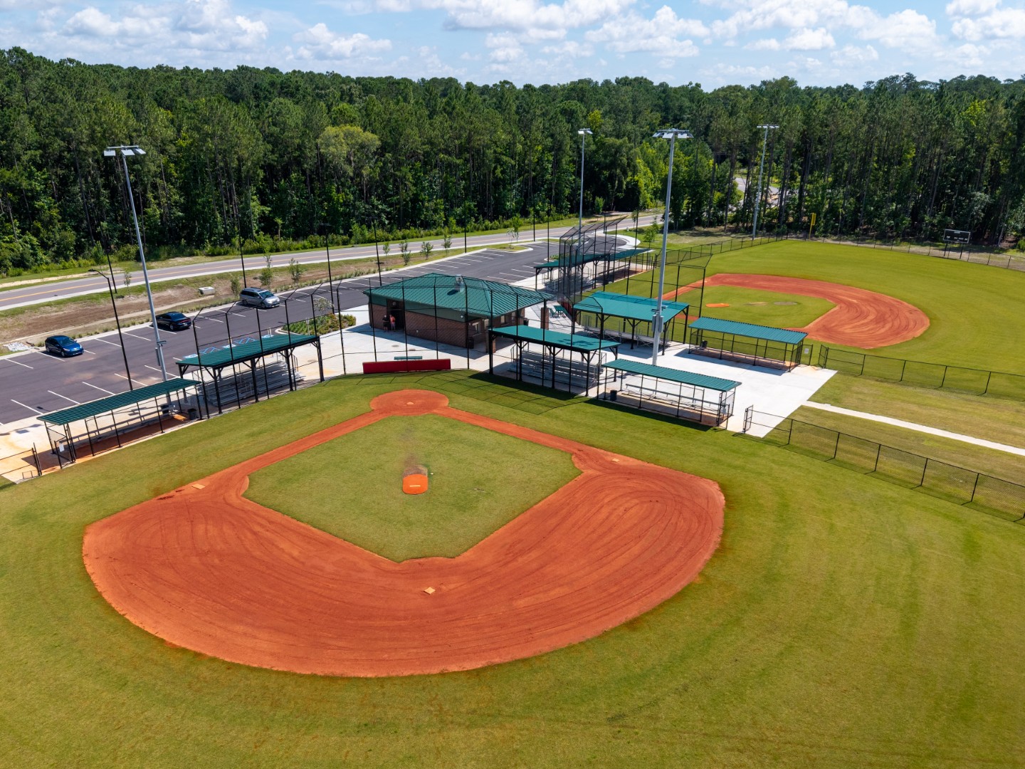 Aerial view of a baseball complex with two red clay fields, green grass, parking lot with cars, and surrounding trees under a partly cloudy sky.