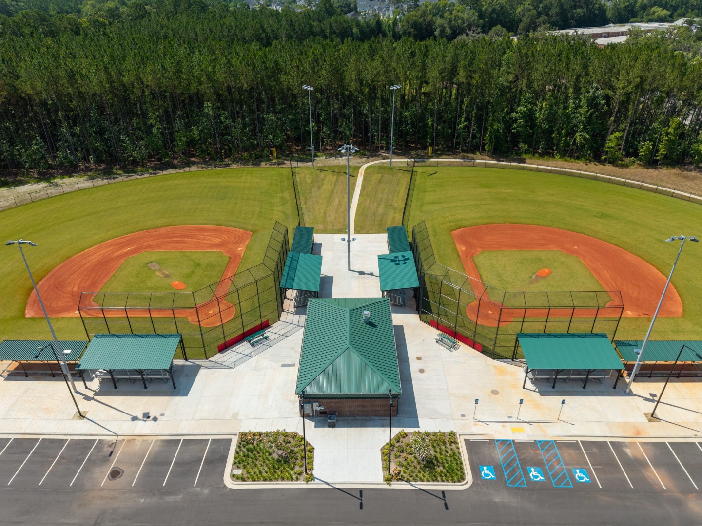 Aerial view of two adjacent baseball fields with red dirt infields, green grass, and fencing. A building with a green roof sits between them, and nearby are benches, walkways, parking spaces, and wooded trees in the background.