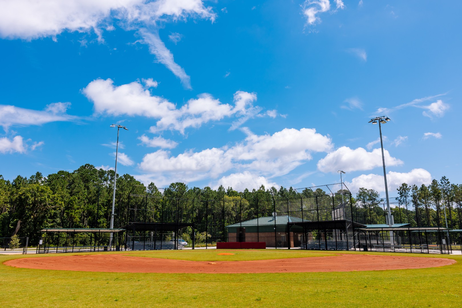 An empty baseball field with a well-kept grass outfield and red dirt infield under a blue sky with scattered clouds, surrounded by trees and a building in the background.