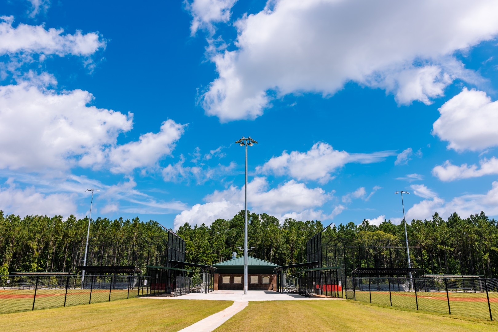 A baseball complex with multiple fenced fields, a central building, and tall light poles, surrounded by green trees under a partly cloudy blue sky.
