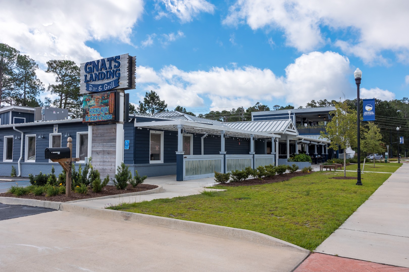 A blue and white building with a sign reading “GNAT’S LANDING Bar & Grill,” surrounded by greenery, a sidewalk, and a partly cloudy sky. The restaurant has a large covered patio and landscaped area in front.