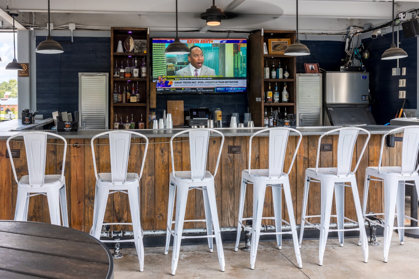 Outdoor bar with five empty white metal stools lined up at a wooden counter. Bottles and glasses are on shelves behind the bar, and a TV above the bar displays a news broadcast. Ceiling fan and lights hang overhead.