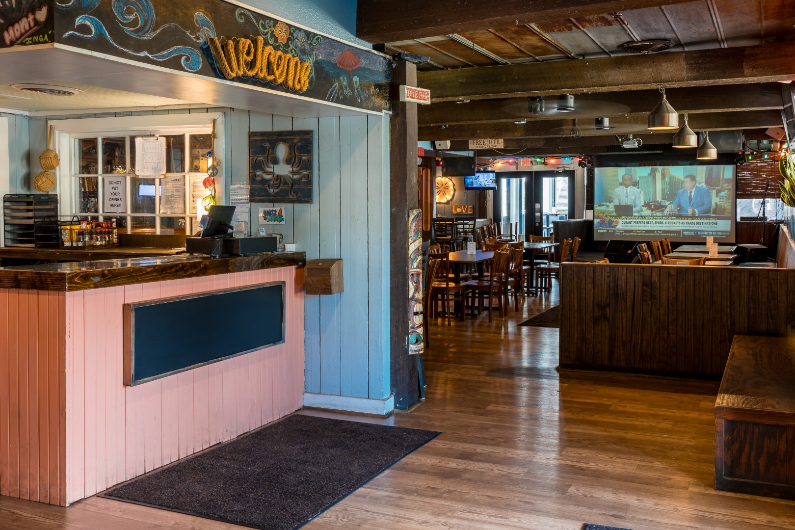 Cozy restaurant interior with a welcome sign above a front counter, wooden floors and furniture, and a dining area in the background with a large screen displaying a news program. Warm lighting creates an inviting atmosphere.