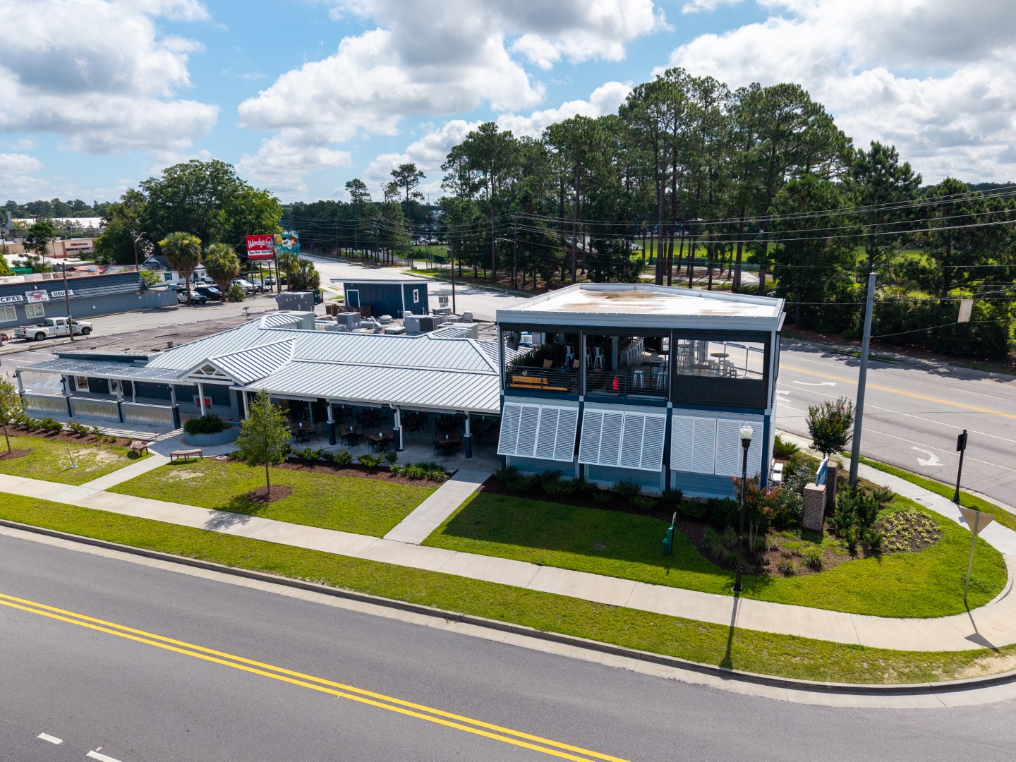Aerial view of a modern restaurant with a rooftop patio, located at a street corner, surrounded by green lawns, trees, and nearby roads under a partly cloudy sky.