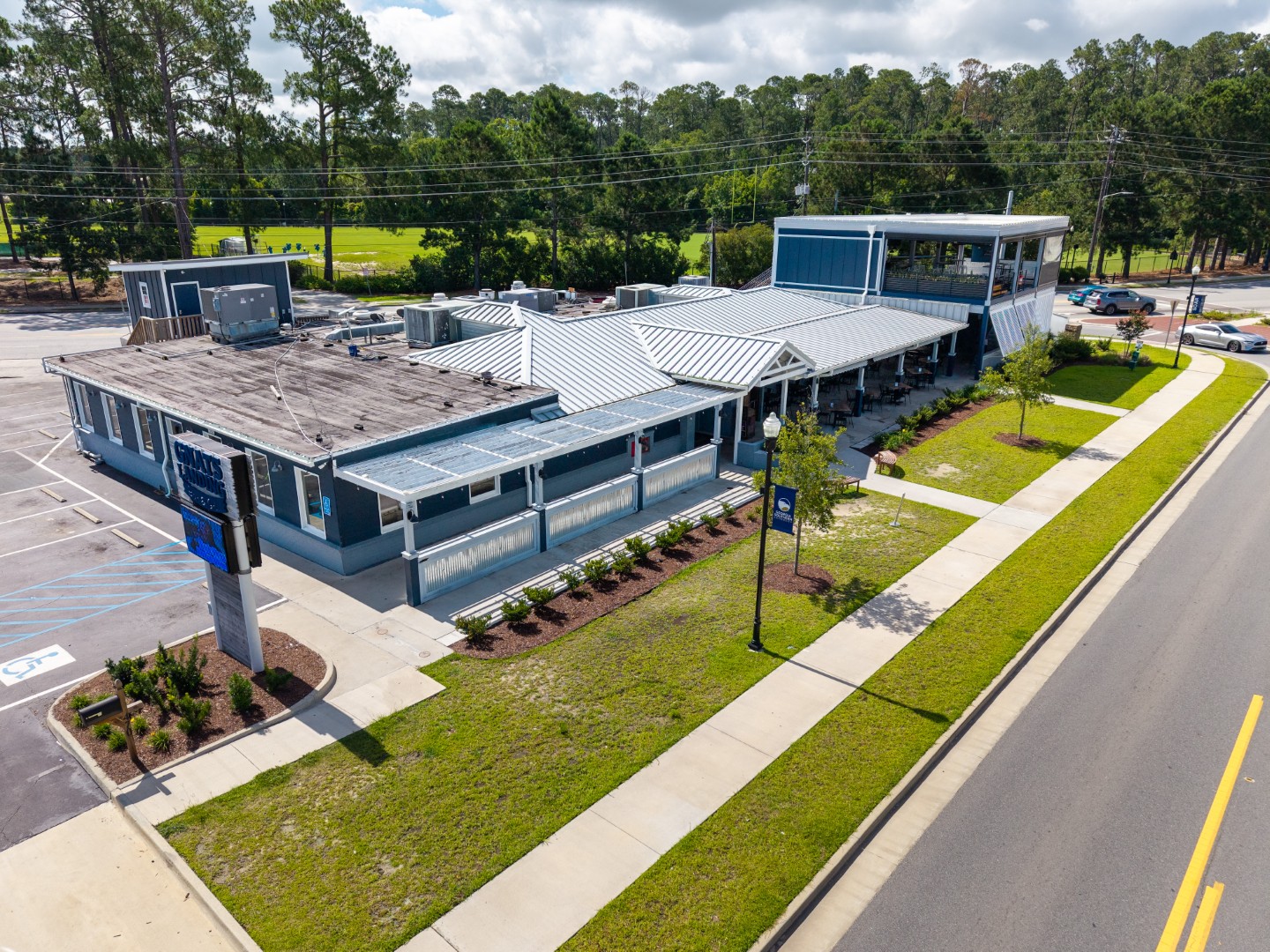 Aerial view of a modern, single-story building with a metal roof, surrounded by trees, parking areas, and well-maintained landscaping along a main road. The scene is bright and partly cloudy.