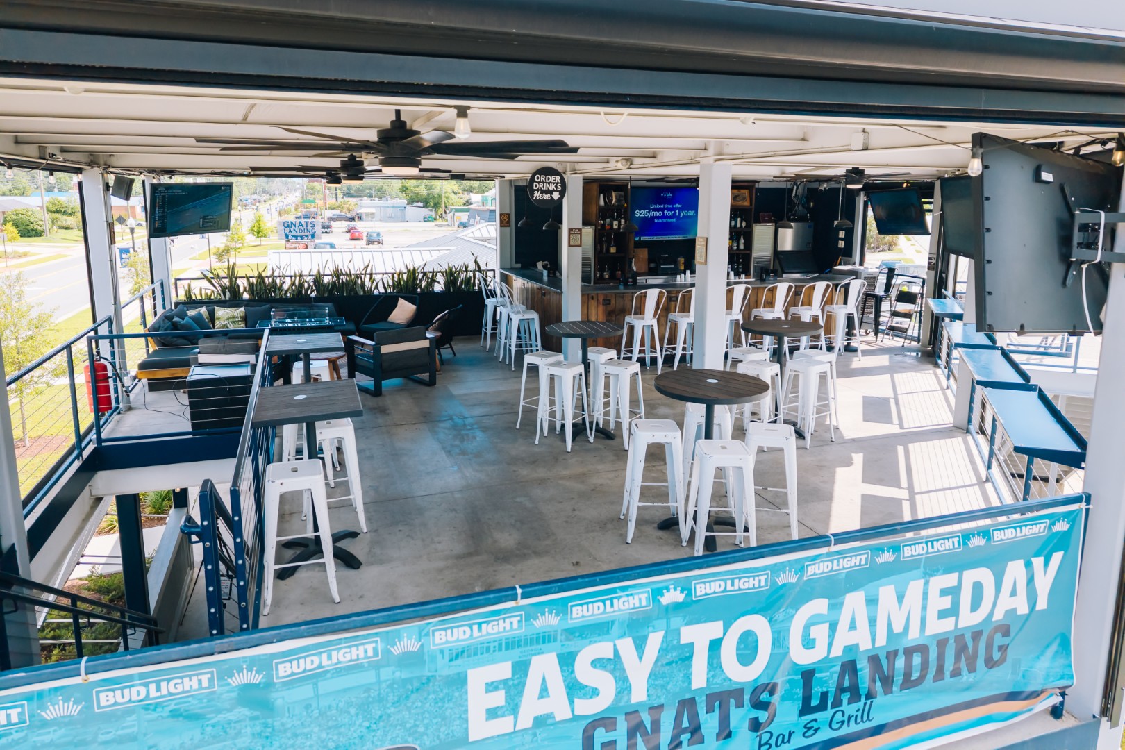 Open-air bar and grill with high tables, white stools, couches, and flat-screen TVs. A Bud Light banner reading “EASY TO GAMEDAY GNAT’S LANDING” hangs on the railing at the front.