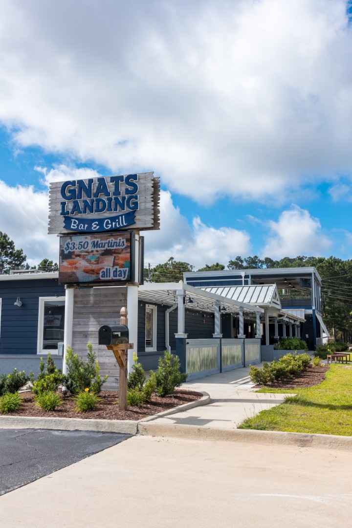 A blue and white building with a sign reading “GNAT’S LANDING Bar & Grill,” surrounded by greenery, a sidewalk, and a partly cloudy sky. The restaurant has a large covered patio and landscaped area in front.