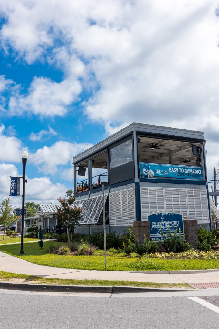 A modern two-story building with a covered outdoor upper deck sits on a corner under a partly cloudy sky. A streetlamp and a blue Soar banner are visible nearby, surrounded by greenery and a sidewalk.