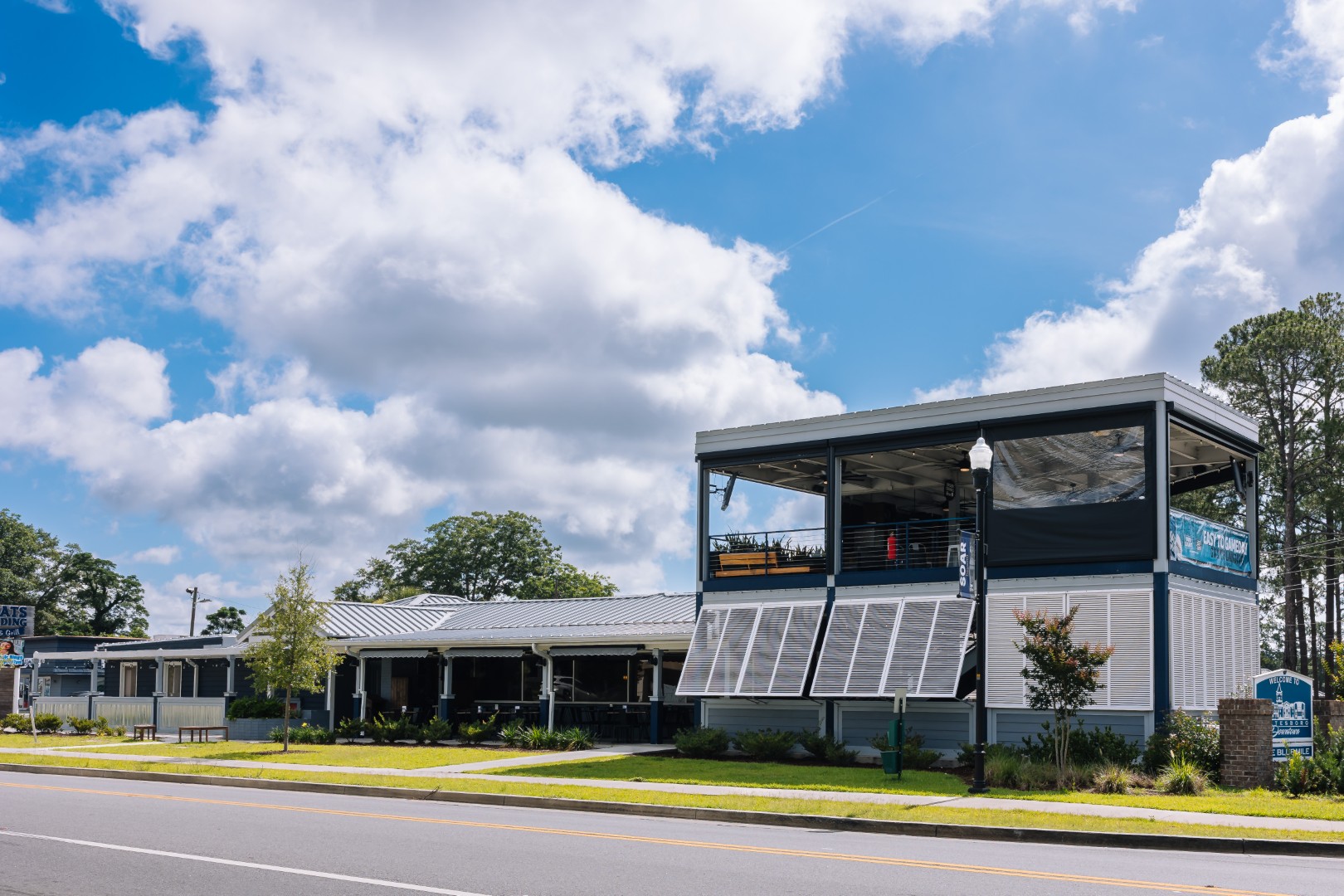 A two-story restaurant with large windows and an outdoor seating area sits along a road on a sunny day with blue skies and scattered clouds. Trees and neatly trimmed grass surround the building.