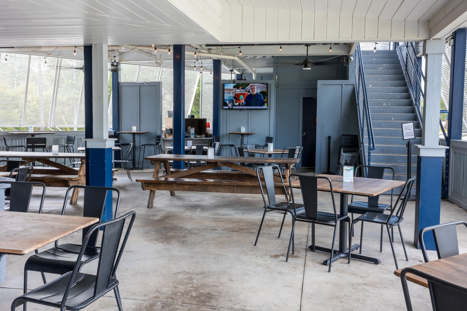 Open-air dining area with wooden picnic tables and black metal chairs, blue and white walls, a staircase, and a TV mounted on the wall showing two people. The space is empty and well-lit.