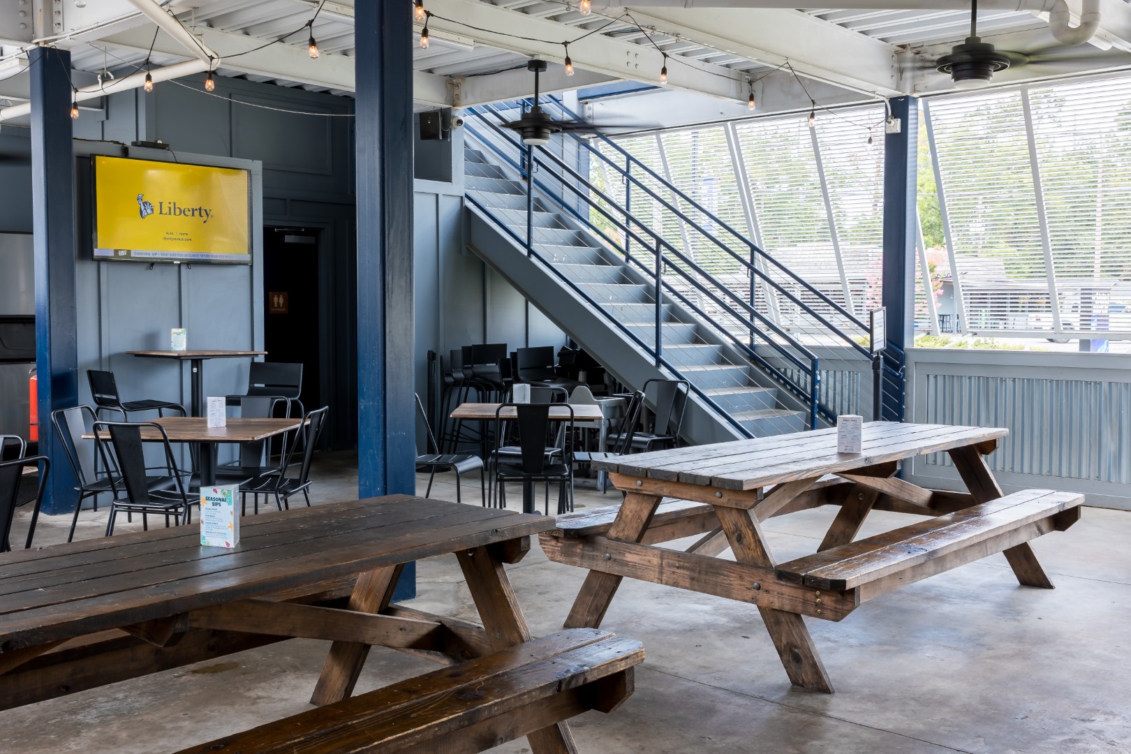 Indoor seating area with wooden picnic tables and metal chairs, large windows letting in natural light, staircase leading to an upper level, and a wall-mounted TV displaying a yellow screen on the left.