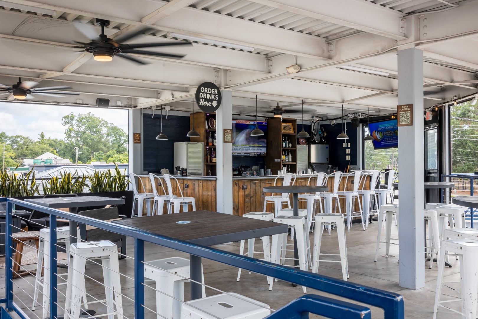 Covered outdoor bar and dining area with high white chairs, tables, ceiling fans, and a wooden bar. TVs are mounted on walls, plants line the railing, and a sign reads “Order Drinks Here.”