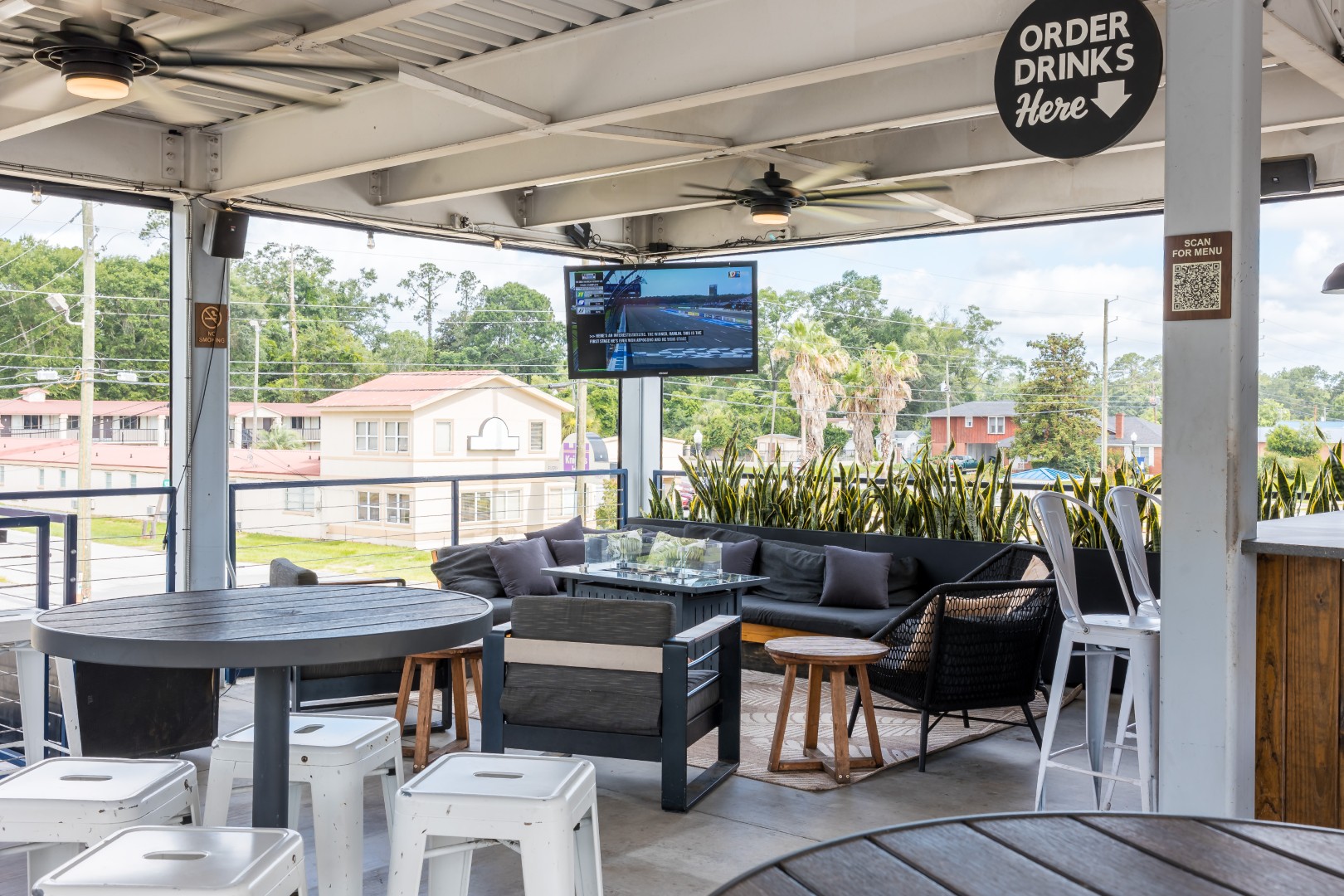 A covered outdoor patio features a TV, round tables with white stools, a black wicker sofa, and potted plants. A sign reads ORDER DRINKS Here. The space overlooks a street with trees and buildings in the background.