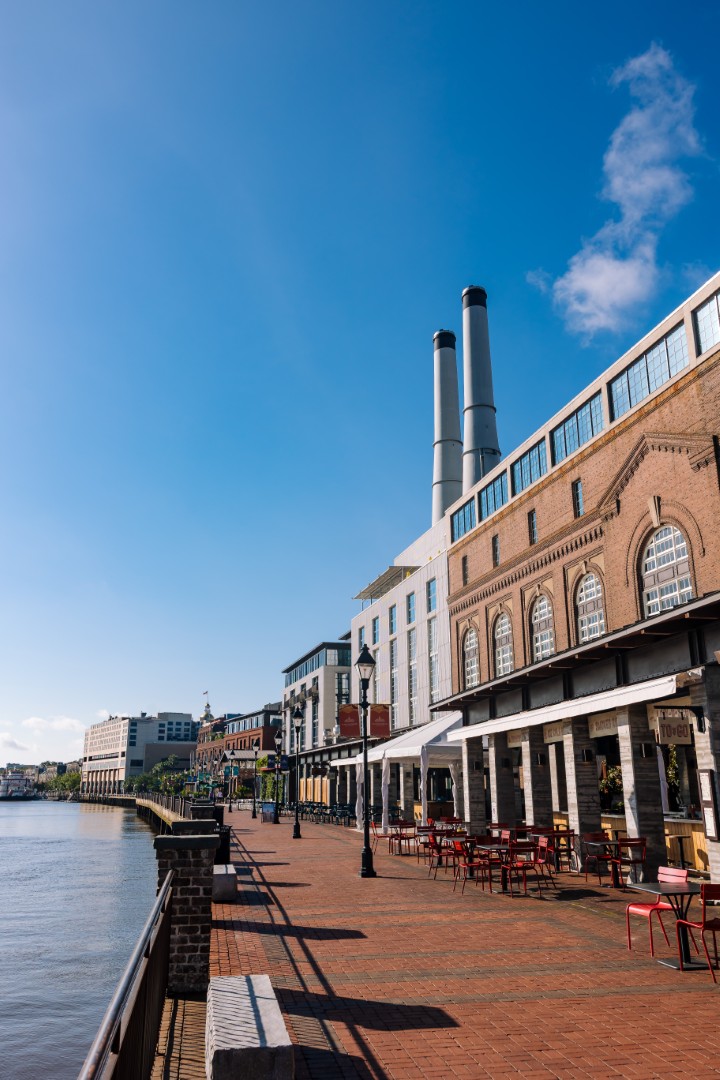 A sunny riverside promenade with red brick walkway, outdoor seating, historic brick buildings, and two tall smokestacks under a clear blue sky.