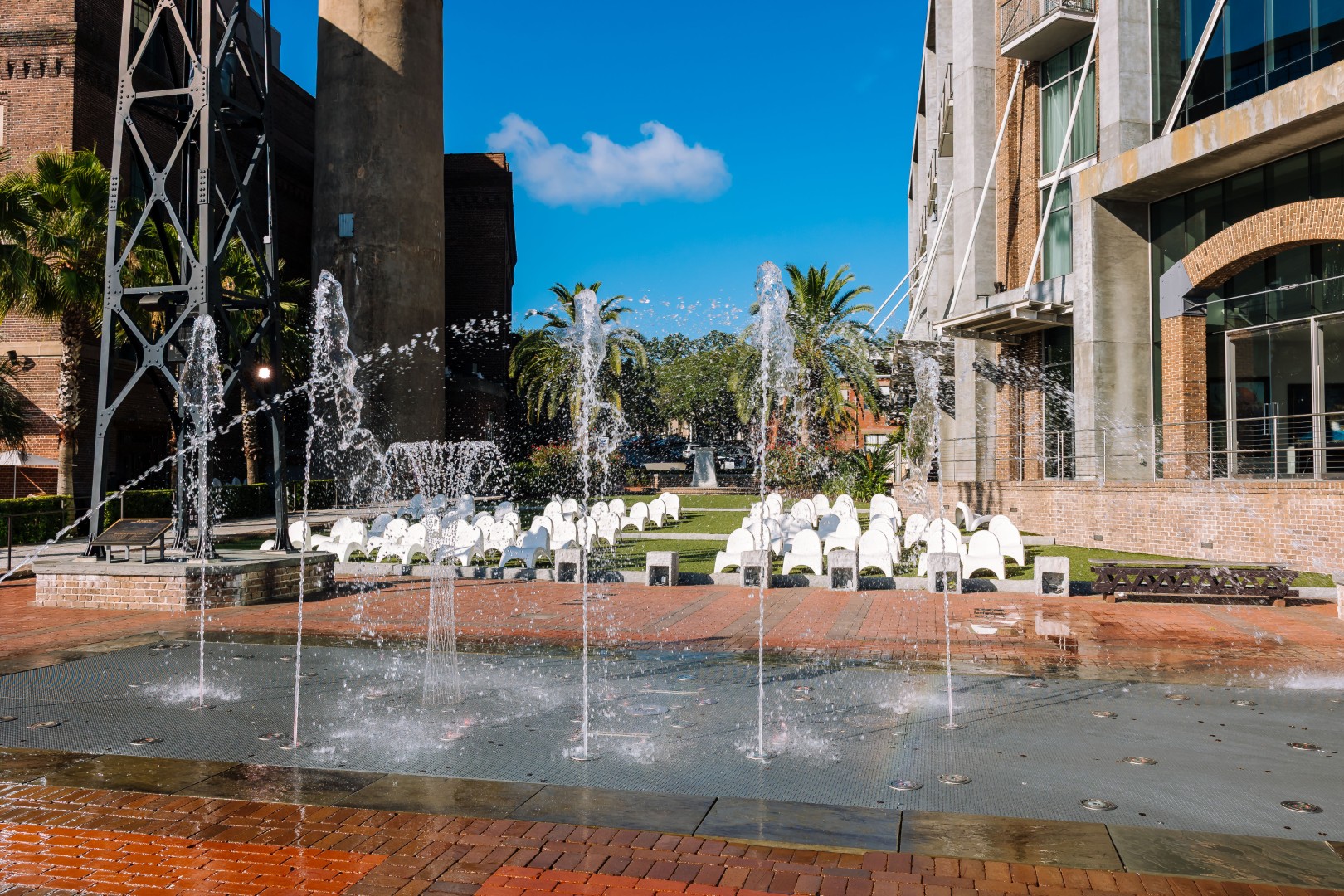 A plaza with water fountains shooting up from the ground, rows of white chairs set up on a lawn, palm trees, and modern buildings under a clear blue sky.