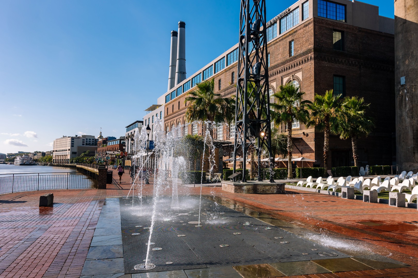 A brick plaza with water fountains, palm trees, and a line of white chairs sits beside a large historic building near a waterfront under a clear blue sky.