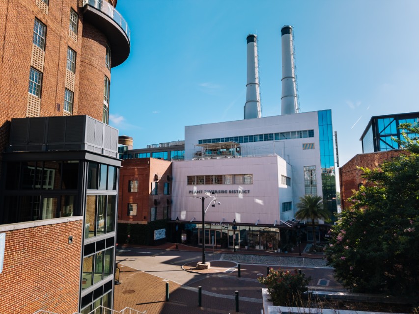 White building with two tall smokestacks stands behind older brick buildings on a sunny day, with a clear blue sky and tree-lined, empty street in the foreground.