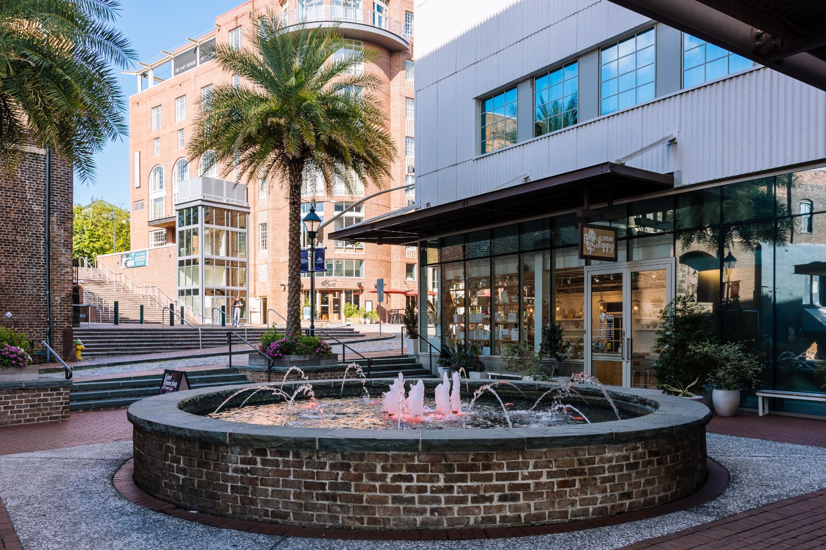 A circular brick fountain with water jets is in a sunny plaza, surrounded by palm trees, potted plants, and modern brick and glass buildings with shops and outdoor steps in the background.