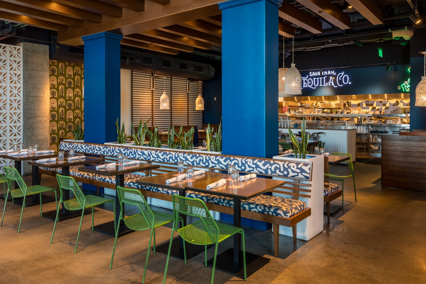 A restaurant interior with blue columns, patterned bench seating, green chairs, set tables, and a neon sign reading Tequila Co. near an open kitchen.