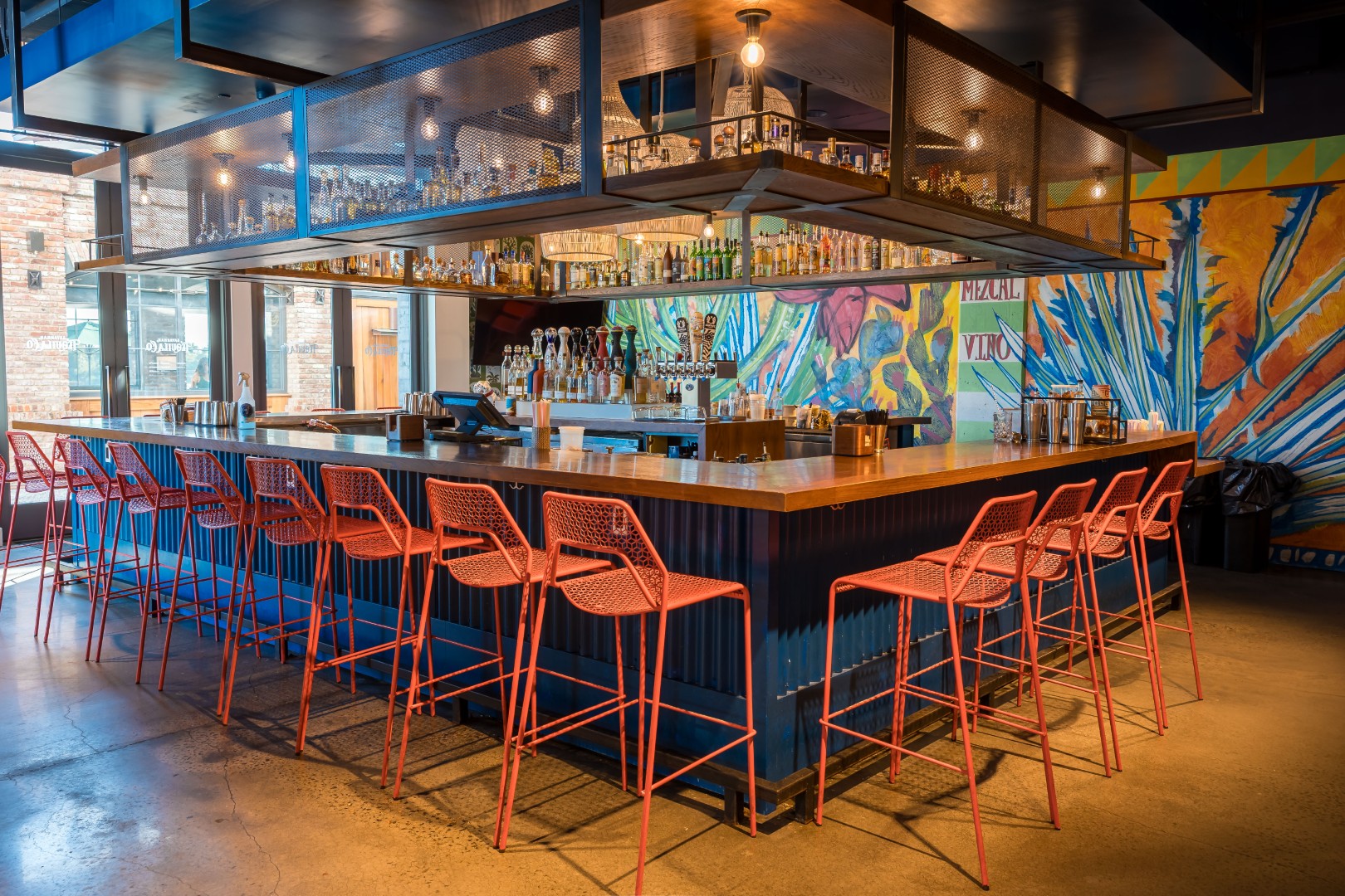A bar with red metal stools lined up along a wooden counter. Colorful mural art covers the walls, and shelves with bottles hang above the bar.