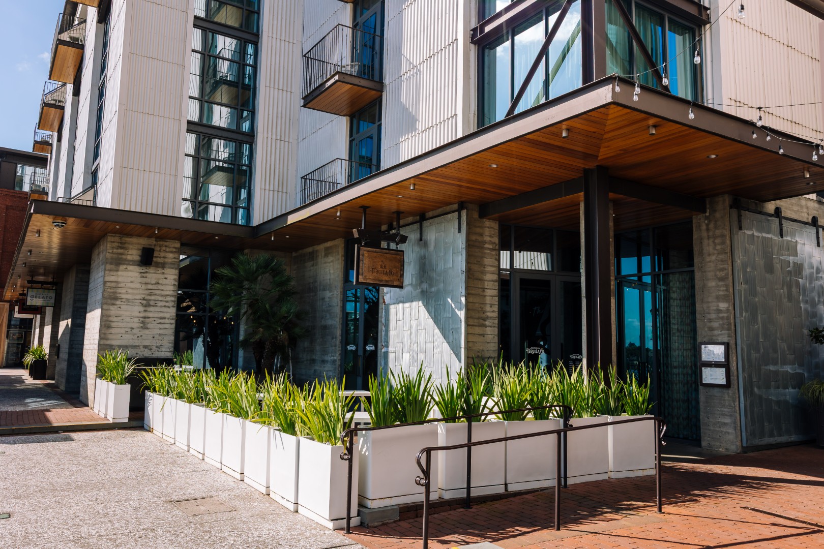 Building with large windows and balconies, featuring a patio area lined with white planters filled with green plants. The entrance has wooden accents and string lights above.
