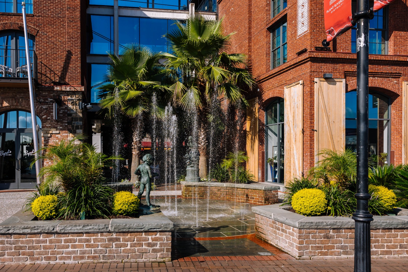 A brick building with large windows stands behind a fountain featuring a bronze statue of a child. Palm trees and green shrubs decorate the fountain area, which is bordered by stone and brickwork.