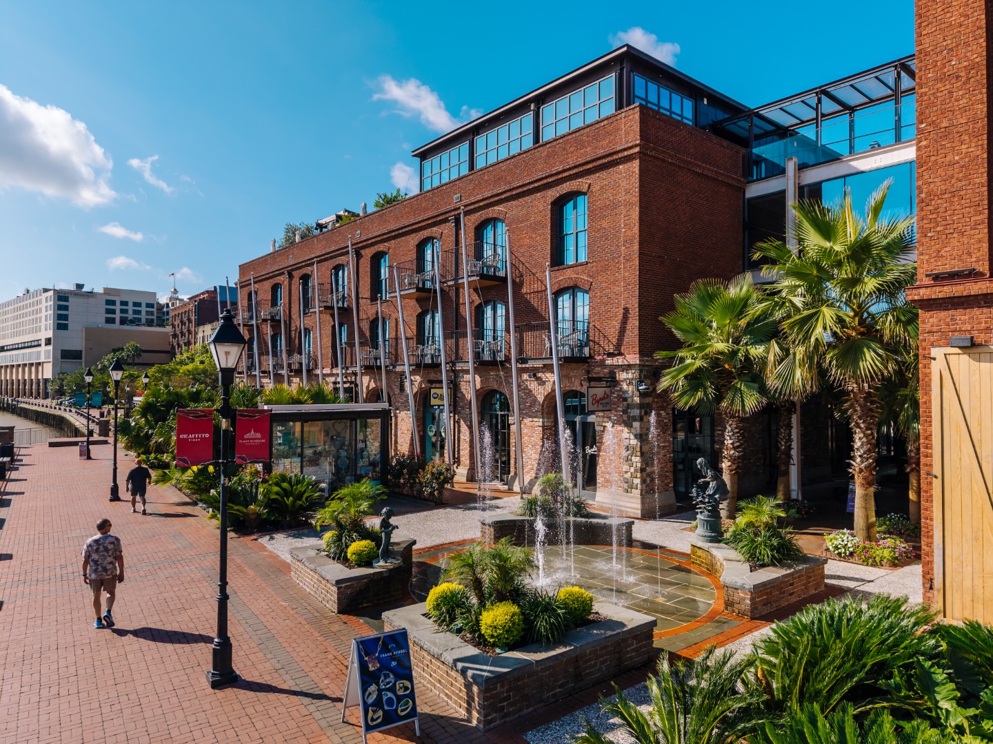 A brick building with balconies and large windows stands along a cobblestone walkway lined with palm trees, greenery, and a small fountain. A few people walk near the shops on a bright, sunny day.